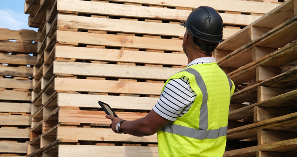 Man wearing a safety vest while holding a tablet looking at a stack of new wooden pallets.