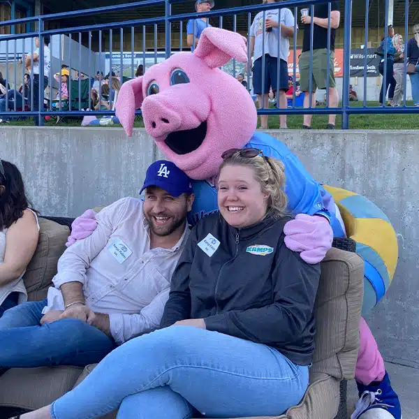 Man and woman laughing while being hugged by mascot at baseball game