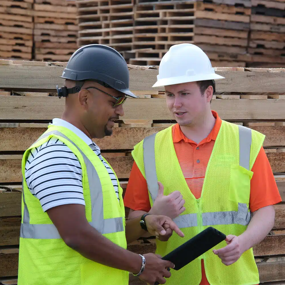 Two men wearing hard hats surrounded by pallets looking at a tablet.