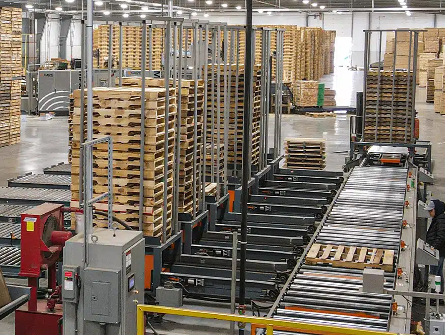 Indoor overhead drone perspective of a pallet manufacturing facility with stacks of new pallets in the background, bright lights, and automation equipment showing a used wooden pallet on a conveyor.