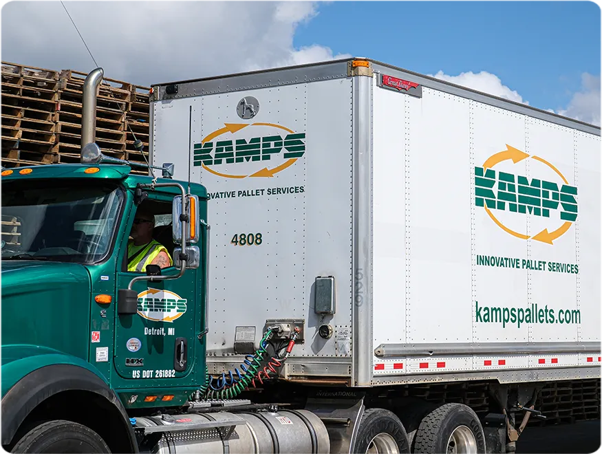 Close up of a green truck and white trailer parked in front of a stack of used wooden pallets